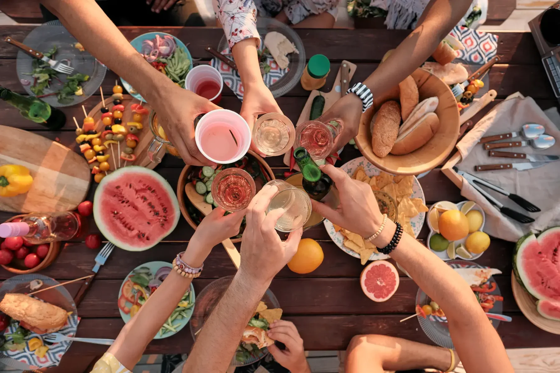A picnic table set with lots of food, people clinking glasses together for a toast